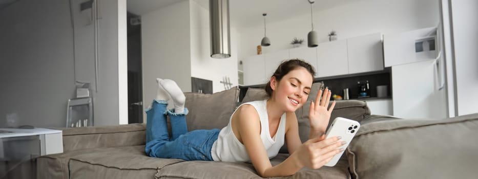 Happy cute female model, lying on couch, waving hand at smartphone, saying hi, connects to video call, resting at home and talking to friend.