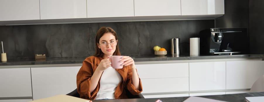 Beautiful young woman having a break from studying, drinking coffee, surrounded by papers and homework, sitting in kitchen at home.