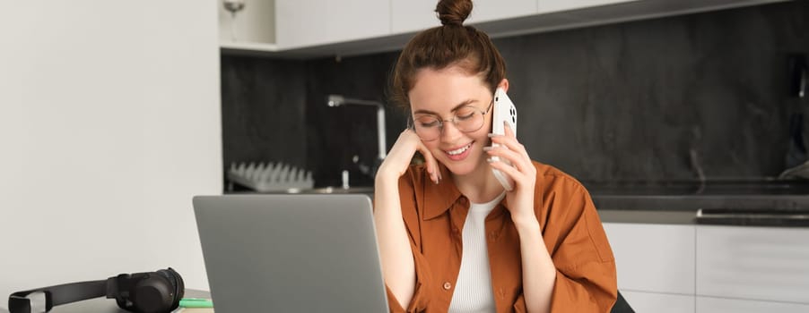 Portrait of young self-employed woman, entrepreneur working from home, freelancer calling client. Girl making an order, talking to someone on phone, sitting in kitchen with laptop.