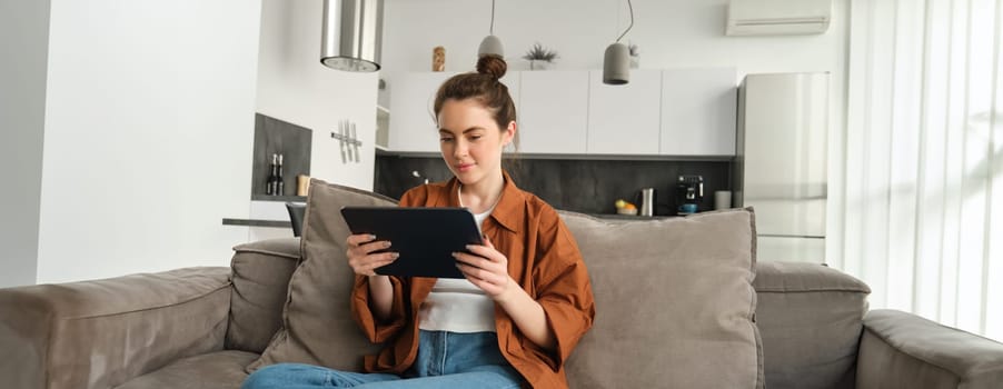 Portrait of young woman sitting on couch in living room, watching movies on digital tablet, reading on device, enjoying weekend at home.