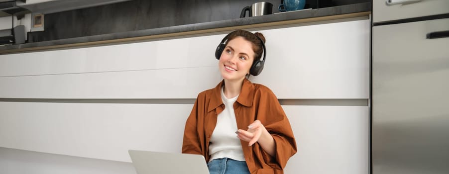 Portrait of young smiling woman explains something over the video call, sits in headphones with laptop, works from home.