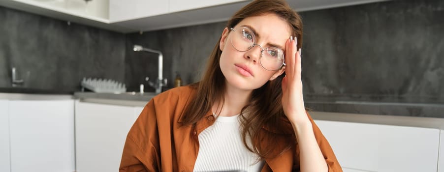 Portrait of brunette woman working from home, wearing glasses, looking concerned, touching head and frowning, holding digital tablet, has headache or migraine.