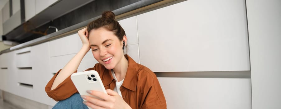 Carefree smiling woman with smartphone, messaging, using mobile phone, sitting on kitchen floor, listening music in wireless headphones.