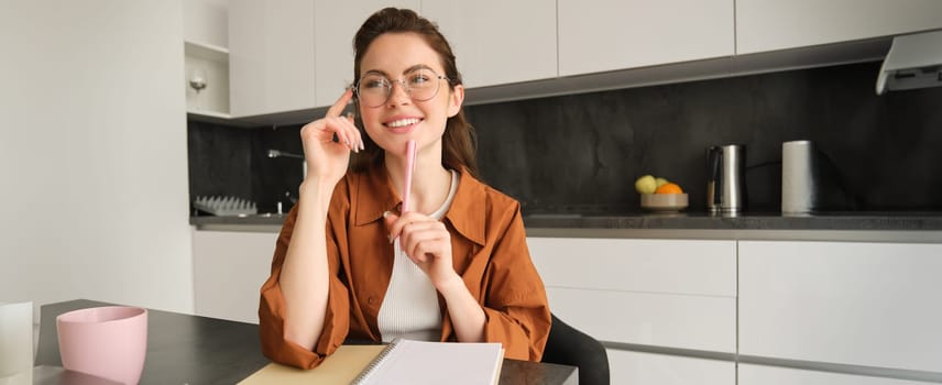Portrait of young creative woman in glasses, student sitting in kitchen, holding pen, writing down information, doing homework, working on project, smiling happy and looking away.