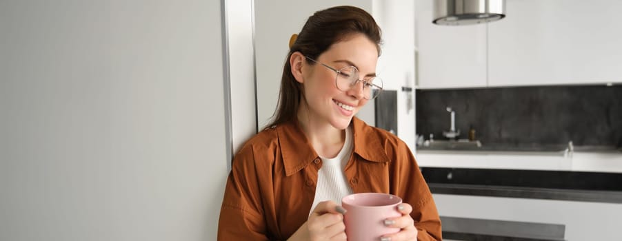 Portrait of modern woman at home, relaxing in her living room, leaning on wall, holding cup of tea and smiling, wearing glasses, drinking coffee and resting.