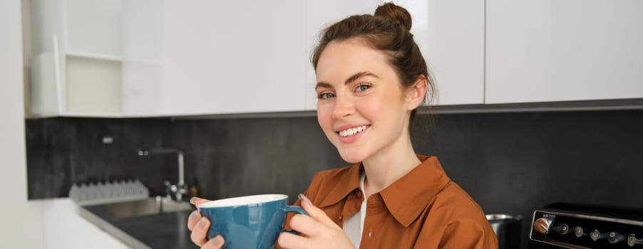 Portrait of happy smiling woman on modern kitchen, drinking aromatic coffee, enjoying freshly made cappuccino at home.