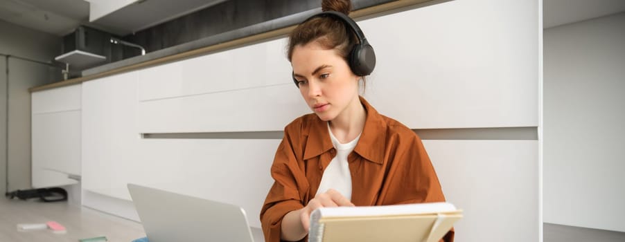 Serious-looking girl student studying at home, sits with laptop and notebook on floor, doing homework project on computer, has online course lesson, wears headphones.