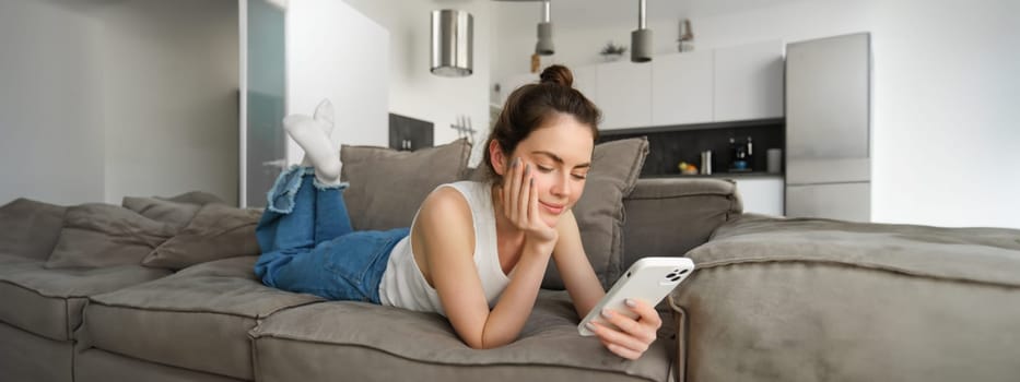 Portrait of cute young woman with smartphone, lying on couch and resting at home, scrolling social media feed, online shopping, reading something on mobile phone.