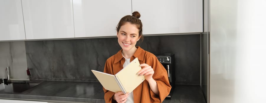 Portrait of beautiful woman standing in the kitchen with notebook, reading recipes, flipping pages and smiling.