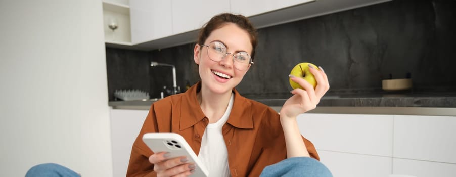 Portrait of happy, beautiful young woman in glasses, sitting with mobile phone, eating apple, laughing and smiling, relaxing, browsing social media application.