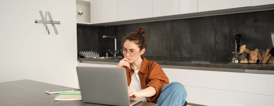 Portrait of woman sitting at home with laptop. Businesswoman managing her own business remotely from her kitchen. Student looking at computer screen, studying, doing her homework.