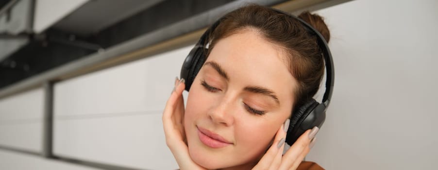 Image of modern woman, beautiful girl enjoys listening to music, has black wireless headphones on, sits on floor and rests.