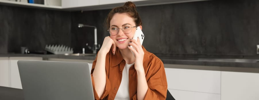 Portrait of beautiful smiling woman working from home, talking on mobile phone, calling client, self-employed businesswoman sets up workplace in kitchen, using laptop and smartphone.