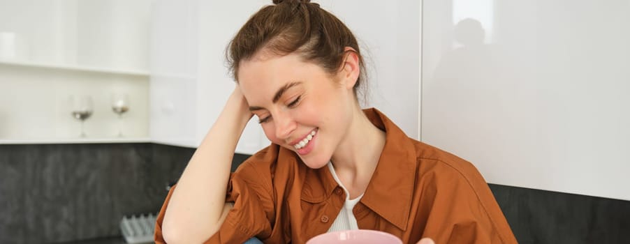 Cosy moments. Happy young woman sits in kitchen and drinks coffee, has tender smile on her face.