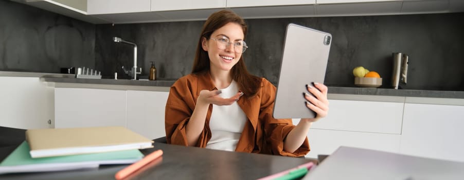 Portrait of young woman working from home, sitting in kitchen and talking with team, has online meeting, using digital tablet to join webinar, having conversation.