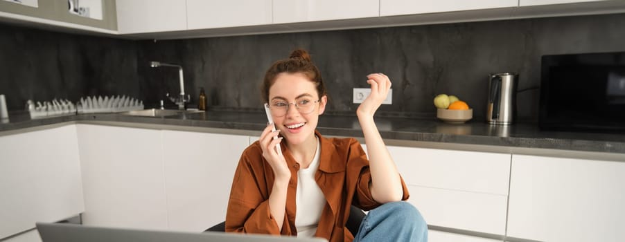 Excited young woman talking on mobile phone in front of laptop, sitting in kitchen with happy face expression, having a conversation.