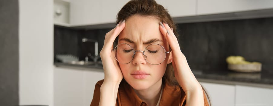 Portrait of tired young working woman in glasses, holds hands on head, suffers painful migraine, has pain in temples, sitting with headache in kitchen.