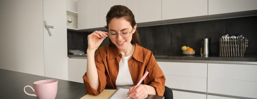 Portrait of young woman student, girl studying at home, working remote in kitchen, taking notes, sitting indoors.