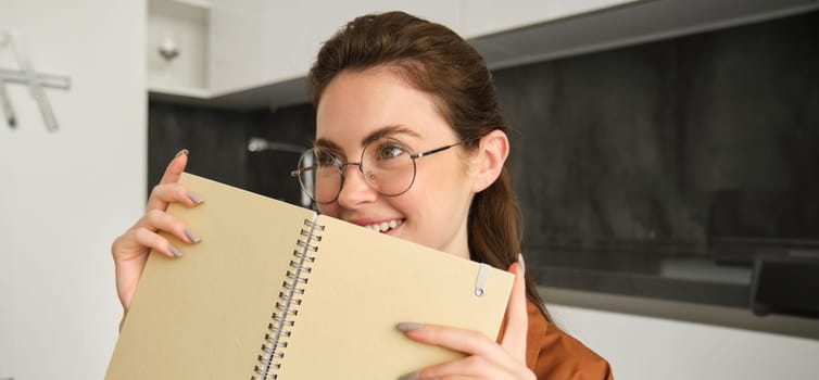 Portrait of beautiful young woman studying, student revising at home, doing homework, holding planner, reading her notes and smiling, writing in diary.