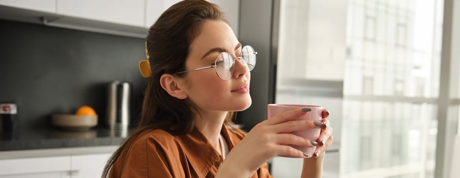 Close up portrait of young beautiful woman in glasses, spending time at home, sitting with cup of coffee, warming up with fresh mug of tea, resting in kitchen.