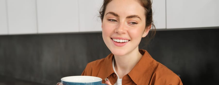 Portrait of smiling beautiful woman at home, drinking coffee, enjoys delicious taste of freshly brewed cappuccino, standing in the kitchen with blue mug.