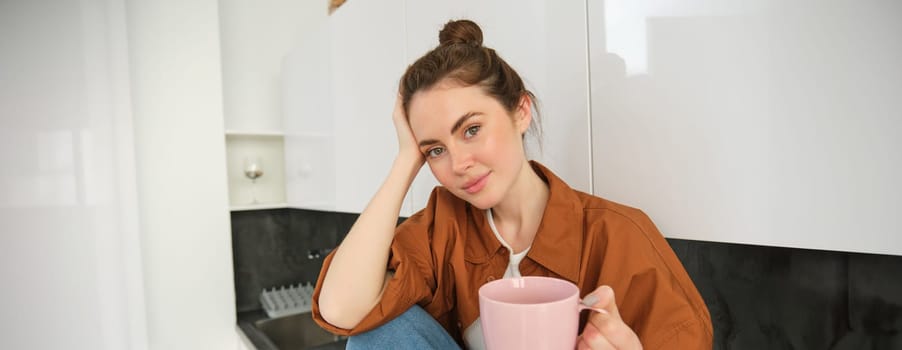 Portrait of young woman with cup of coffee, sits in kitchen and drinks aromatic drink at home, holds tea mug.