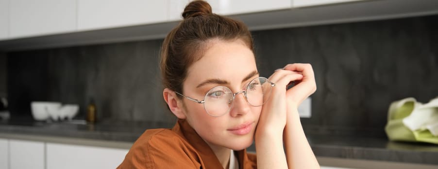 Close up portrait of gorgeous young woman in glasses, looking thoughtful and relaxed, posing sitting in kitchen, smiling with tenderness.