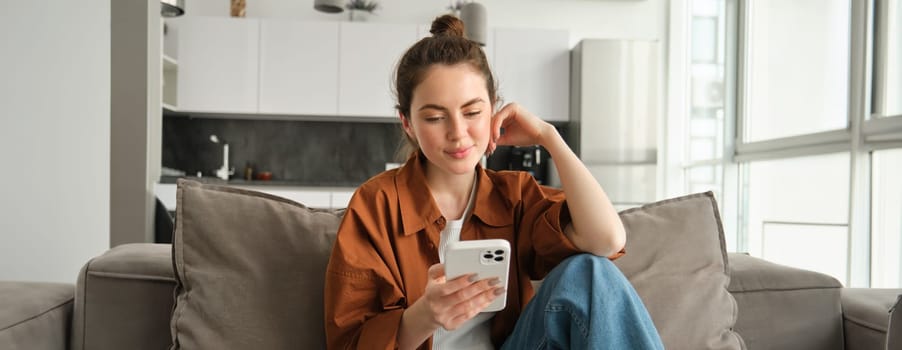 Portrait of happy young brunette woman sitting on sofa with smartphone, reading notification, messaging on mobile phone, resting on couch at home, smiling while looking at device screen.