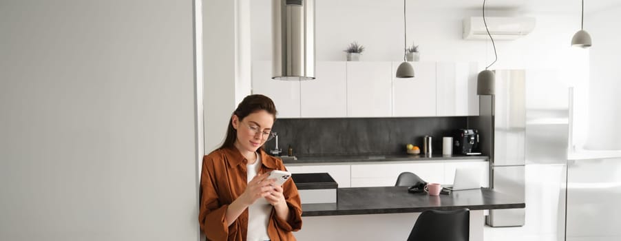 Portrait of young woman at home, checking news or messages on smartphone, online shopping, confirm delivery on mobile phone app, leaning on wall, standing in living room.