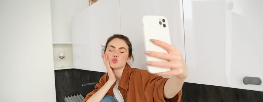 Young charismatic girl takes selfies in the kitchen, posing for photo, holds mobile phone with extended hand.