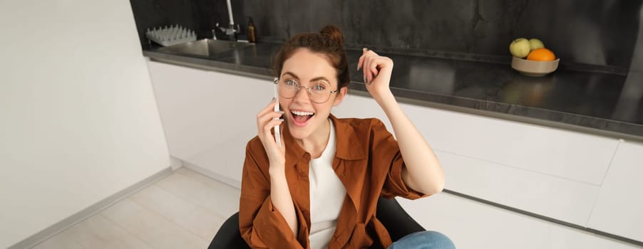 Portrait of woman receiving great news over the phone call, celebrates and jumps on chair from excitement during telephone conversation.