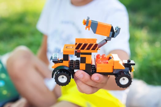 A child s hands holding a toy truck made of building blocks. The truck is orange and black with stripes on the side. The child is sitting on a grassy lawn in a park on a sunny day