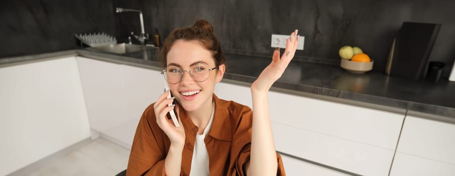 Image of beautiful modern woman working from home, studying in kitchen with laptop, talking on smartphone, calling someone on telephone.