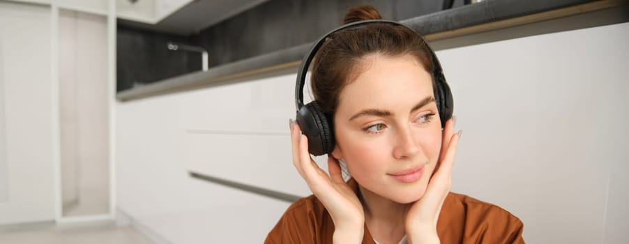 Image of modern woman, beautiful girl enjoys listening to music, has black wireless headphones on, sits on floor and rests.