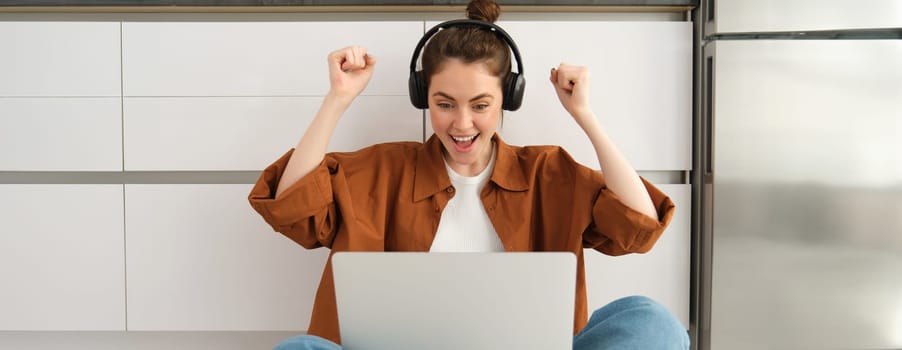 Excited young woman, programmer achieves goal, triumphing, winning on laptop, sitting in headphones, dancing with hands, celebrating victory.