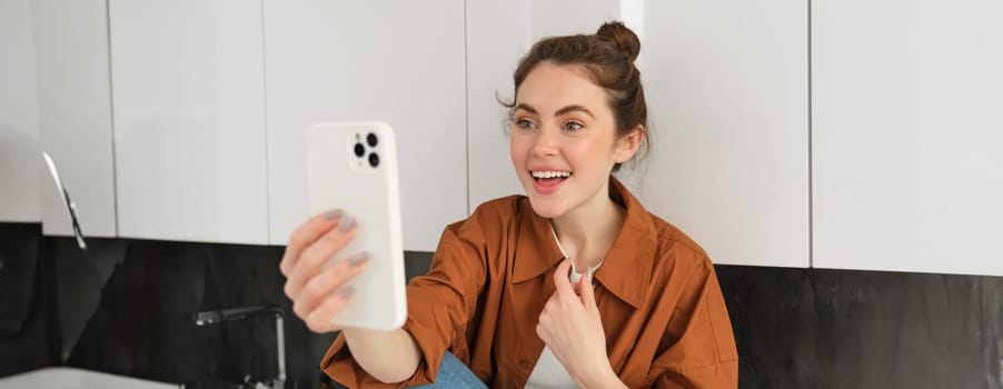 Happy young woman at home, chats with friend on video, holds her smartphone, records lifestyle blog from her kitchen, talking to subscribers on mobile app.
