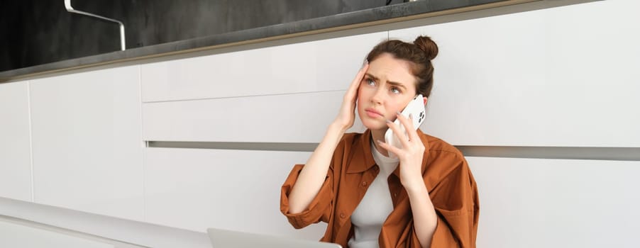 Portrait of woman calling her doctor on the phone, holding smartphone, frowning and touching her head, has painful headache or migraine, feeling unwell.