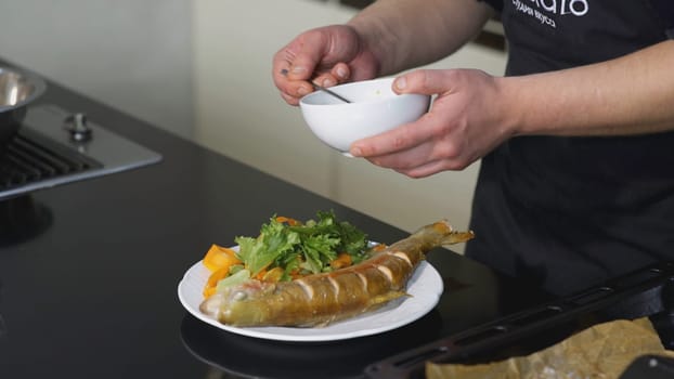 The cook is pouring sauce on a fish dish. ART. Delicious fish dish on a white plate. Professional serving of fish garnished with brown sauce.