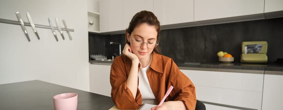 Portrait of young woman, student doing her homework, studying at home, sitting in kitchen, making notes, writing down information.