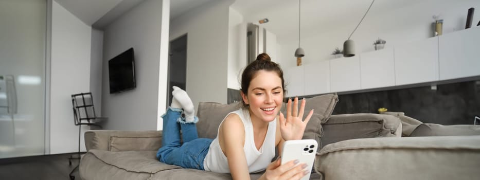 Portrait of happy, smiling young woman online chatting, waving hand at smartphone camera, saying hello while calling someone on mobile phone, spending time in living room.
