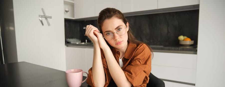 Portrait of young serious woman, working from home in glasses, sitting with folder and documents in kitchen, looking tired.