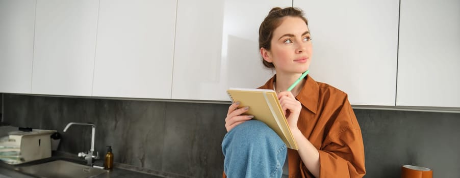 Portrait of modern young woman with notebook and pen, sitting in the kitchen, looking away with thinking face, writing her ideas in planner, creating a wish list.