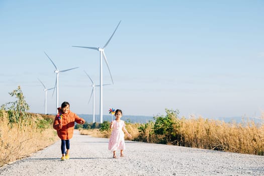 Running girl pinwheel in hand and her sister near windmills. Children embrace turbines signifying sustainable future. Joyful kids windmill in sky symbolize cheerful clean energy-driven community.