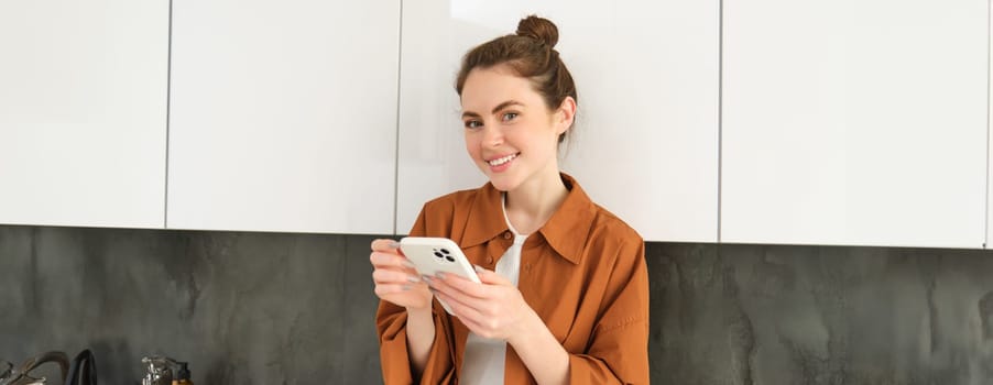 Portrait of young smiling woman with mobile phone, standing in the kitchen, looking happy and relaxed at camera.