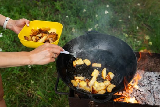 A man cooks potatoes in a wok over an open fire. Place the finished potatoes into a yellow bowl. The background is a grassy field. Outdoor picnic