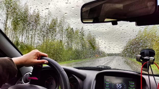 View from car windshield from the driver's seat and hand on the steering wheel of woman on during rain, drops on the glass and nature. Travel in rainy autumn, spring or summer weather