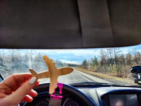 Small wooden toy airplane in the female driver's hand in car on the background of the road and highway. The concept of choosing the type of transport to travel. Choosing between an airplane and car