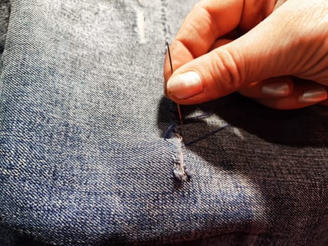 Woman repairing a pair of blue jeans. Fingers with a needle and torn jeans fabric. Sewing up a tear on blue fabric