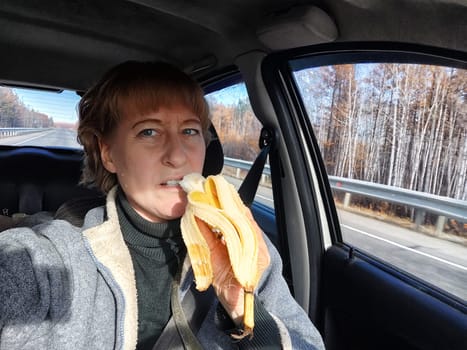 Portrait of female driver in solo journey. Adult mature middle aged woman holding steering wheel and a banana. Eating while driving for to stay awake. Lady girl with food while travel or trip by car