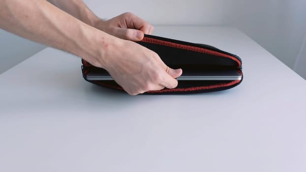The guy putting small silver laptop into a bag and zipping it up on white background of the table and the wall. Transporting a portable computer inside a protective folder.